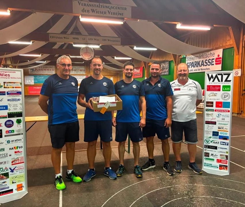 Fünf Männer in passenden blauen und weißen Uniformen stehen im Kreis, einer hält eine Box. Sie befinden sich in einer Sporteinrichtung mit Bannern und einer Uhr an der Wand.