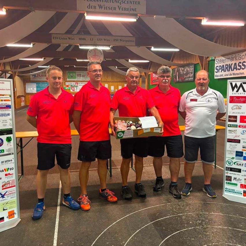 Fünf Männer in roten Shirts und schwarzen Shorts stehen in einer Sporthalle, einer hält eine Box mit Essen. Hinter ihnen steht ein Banner mit der Aufschrift 'Sparkass'.