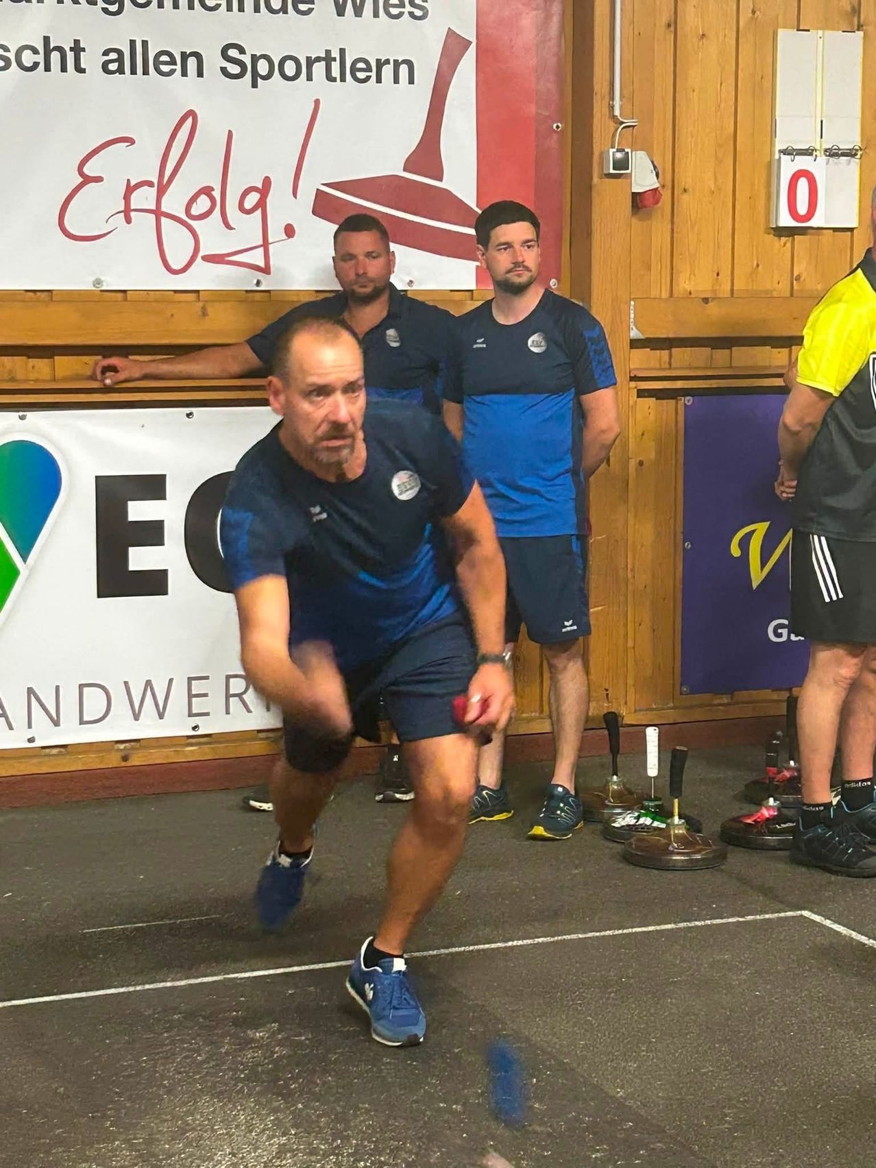 Ein Mann in einem blau-weißen Sportanzug führt einen Kettlebell-Swing in einem Fitnessstudio aus. Drei Männer in blauen Uniformen stehen hinter ihm. Ein Banner mit dem Wort Erfolg ist an der Wand.