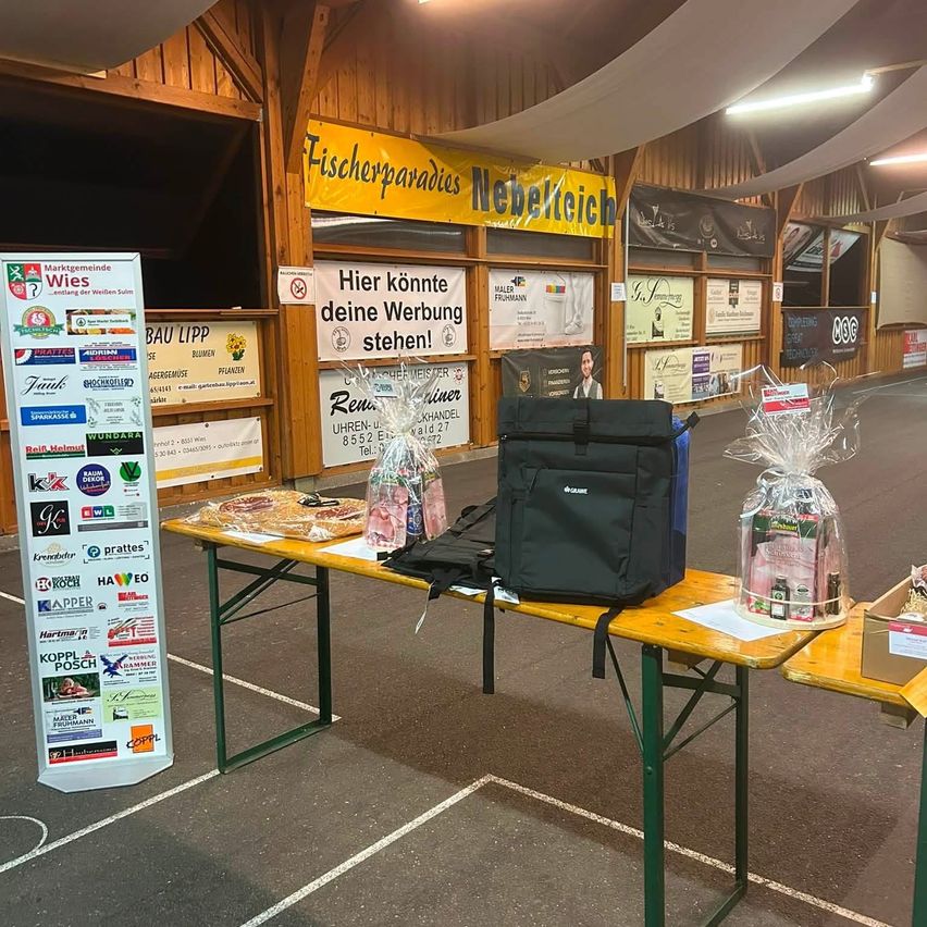 Ein Holztisch mit verschiedenen Gegenständen darauf, einschließlich einer schwarzen Tasche, einem eingewickelten Geschenk und einer Schachtel. Im Hintergrund befinden sich Banner und eine Holzwand.