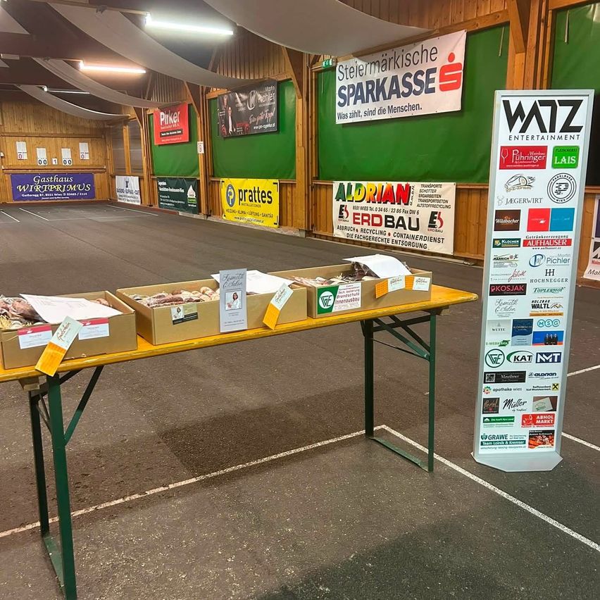 Ein gelber Tisch mit drei Lebensmittelkisten steht in einem großen Innenbereich mit Bannern an den Wänden. Der Tisch steht vor einem weißen Schild mit den Buchstaben WATZ.