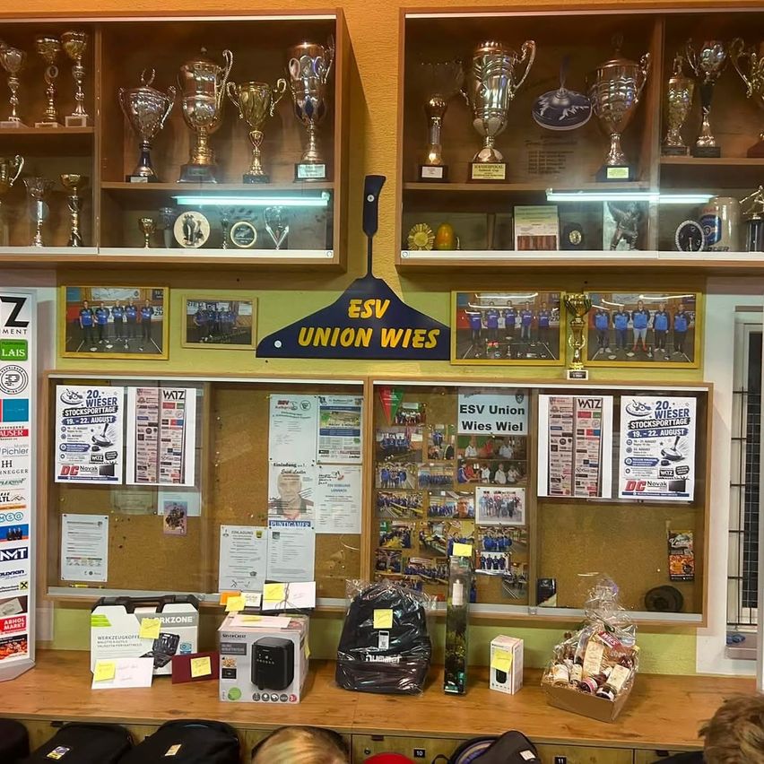 Ein Sportclub-Display mit Trophäen auf Regalen, einer Pinnwand mit verschiedenen Papieren und einem Schreibtisch mit Taschen und einem Korb mit Gegenständen. Die Wand ist gelb gestrichen.