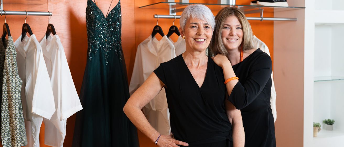 Zwei Frauen stehen in einem Bekleidungsgeschäft zusammen und tragen beide schwarze Kleider. Eine hat ihre Hand auf der Schulter der anderen. Hinter ihnen hängen Kleider an der Wand und Schuhe sind auf einem Regal.