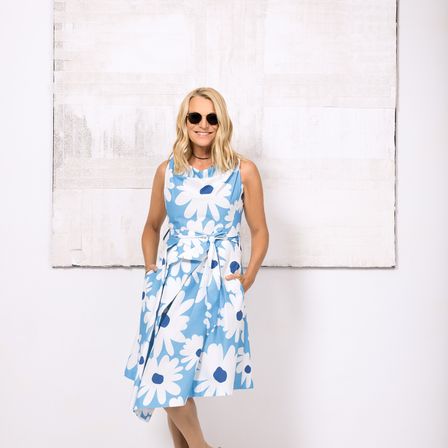 Eine Frau steht vor einer weißen Wand mit einem großen abstrakten Gemälde, trägt ein blau-weißes Blumenkleid und eine Sonnenbrille.