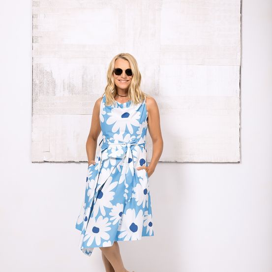 Eine Frau steht vor einer weißen Wand mit einem großen abstrakten Gemälde, trägt ein blau-weißes Blumenkleid und eine Sonnenbrille.