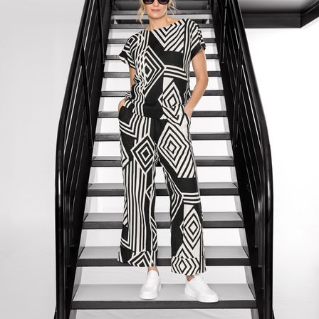 Eine Frau in einem schwarz-weiß gemusterten Jumpsuit steht auf einer modernen Treppe. Sie trägt weiße Sneakers und hat die Hände in den Taschen.