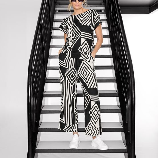 Eine Frau in einem schwarz-weiß gemusterten Jumpsuit steht auf einer modernen Treppe. Sie trägt weiße Sneakers und hat die Hände in den Taschen.