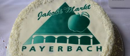 Ein Kuchen mit einem grünen Apfel, einer Brücke und dem Logo Jakobi Markt Payerbach auf weißer Glasur. Er steht auf einem Silberteller mit Tischdecke.