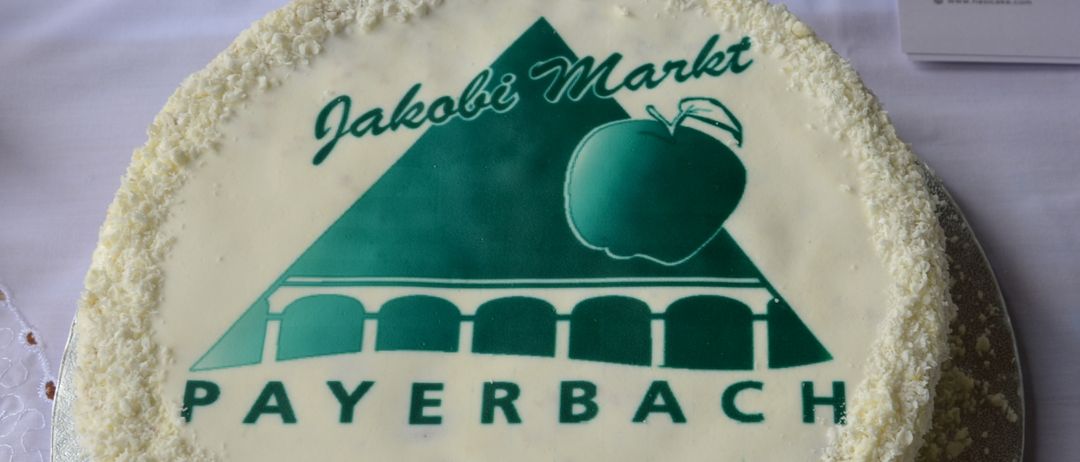 Ein Kuchen mit einem grünen Apfel, einer Brücke und dem Logo Jakobi Markt Payerbach auf weißer Glasur. Er steht auf einem Silberteller mit Tischdecke.
