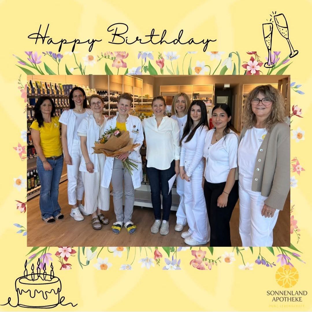 A group of women in white lab coats and glasses pose for a birthday photo in a pharmacy. Flowers and a birthday cake are in the foreground.