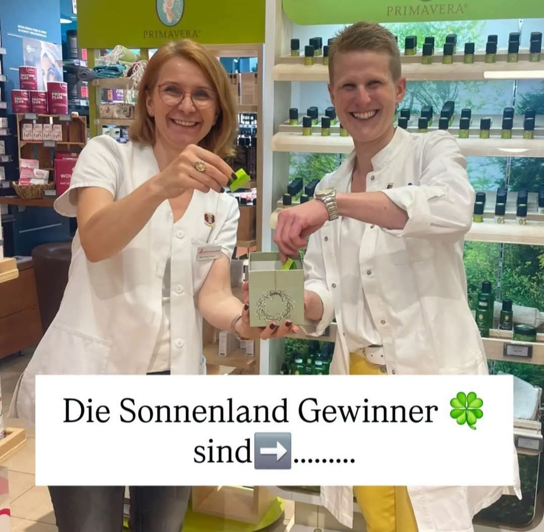 Two people in white lab coats are smiling and holding a small box. They are standing in front of a shelf with various products. The woman is wearing glasses and a ring. The man is wearing a watch. The image has a watermark that reads 'Die Sonnenland Gewinner sind'.