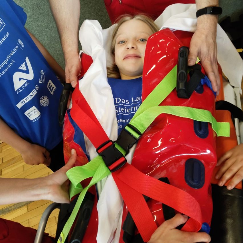 Ein junges Mädchen liegt in einem roten Krankentragen, trägt ein blaues T-Shirt, während Erwachsene ihr helfen. Die Trage hat rote und grüne Gurte und einen schwarzen Griff. Die Umgebung scheint drinnen zu sein, möglicherweise in einem Krankenhaus.