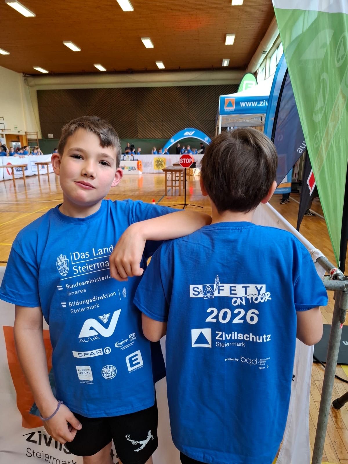 Zwei junge Jungen stehen in einem Innenbereich, einer mit dem Arm um den anderen, beide tragen blaue T-Shirts mit Sicherheitsslogans. Hinter ihnen sind ein Stoppschild und eine Fahne zu sehen.