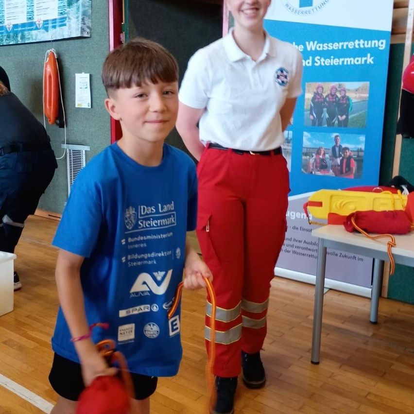 Ein junger Junge und ein Mädchen stehen zusammen, das Mädchen in roten Hosen und einem weißen Shirt, und der Junge in einem blauen Shirt. Sie halten rote Taschen mit Seilen. Hinter ihnen steht ein Tisch mit verschiedenen Gegenständen und einem Banner.
