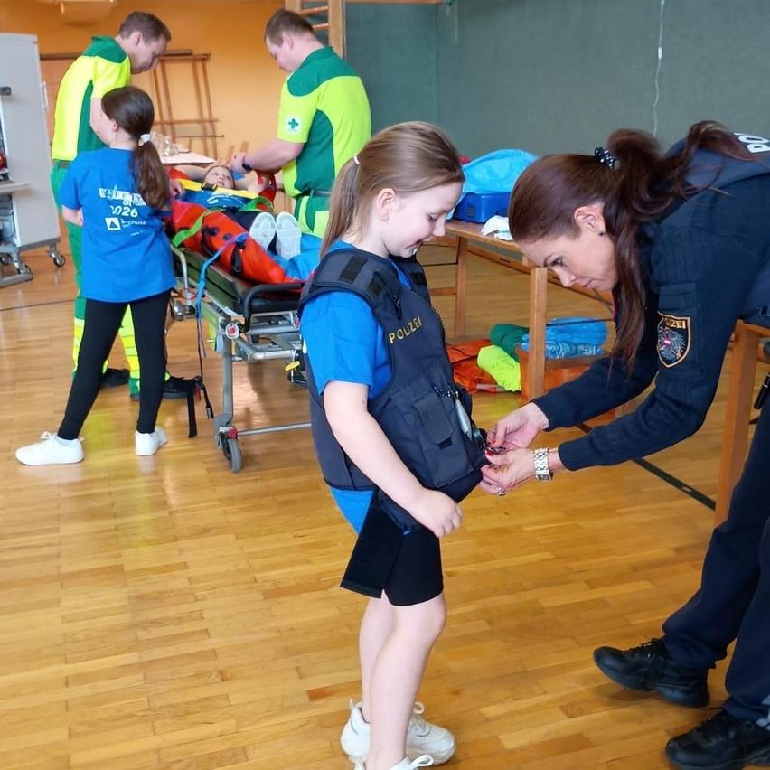 Eine Frau hilft einem Mädchen, einen Schwimmwesten anzulegen. In der Nähe liegt eine Person auf einer Trage, die von zwei Personen in grünen Uniformen betreut wird. Eine weitere Person in einer blauen Uniform beobachtet.