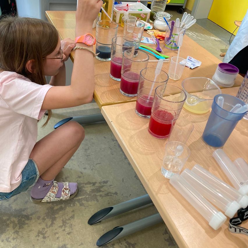 Ein junges Mädchen mit Brille und einem rosa T-Shirt ist an einem wissenschaftlichen Experiment beteiligt und gießt farbige Flüssigkeiten in Gläser auf einem Holztisch.