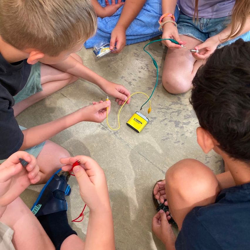 Gruppe von Kindern, die an einem wissenschaftlichen Experiment arbeiten, wobei sie Drähte und ein gelbes Gerät mit der Aufschrift 'UVARTA' verwenden. Sie sitzen auf dem Boden, einige in Sandalen, einige in Shorts, und sehen konzentriert aus.