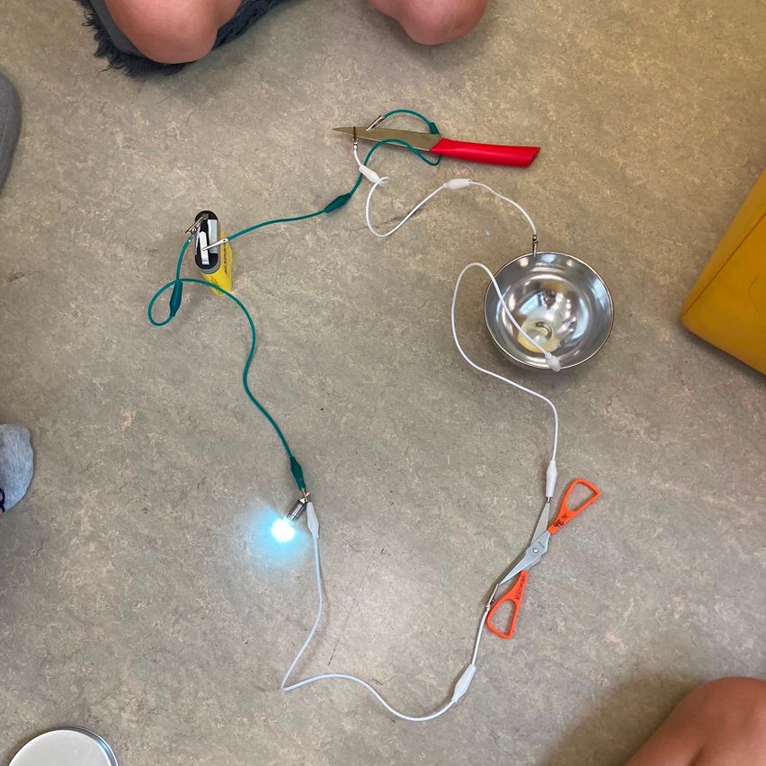 Ein elektrisches Experimentaufbau auf dem Boden mit Drähten, die mit einer Batterie, einer Lampe und einer metallischen Schüssel verbunden sind. Eine Schere und Zangen liegen in der Nähe.