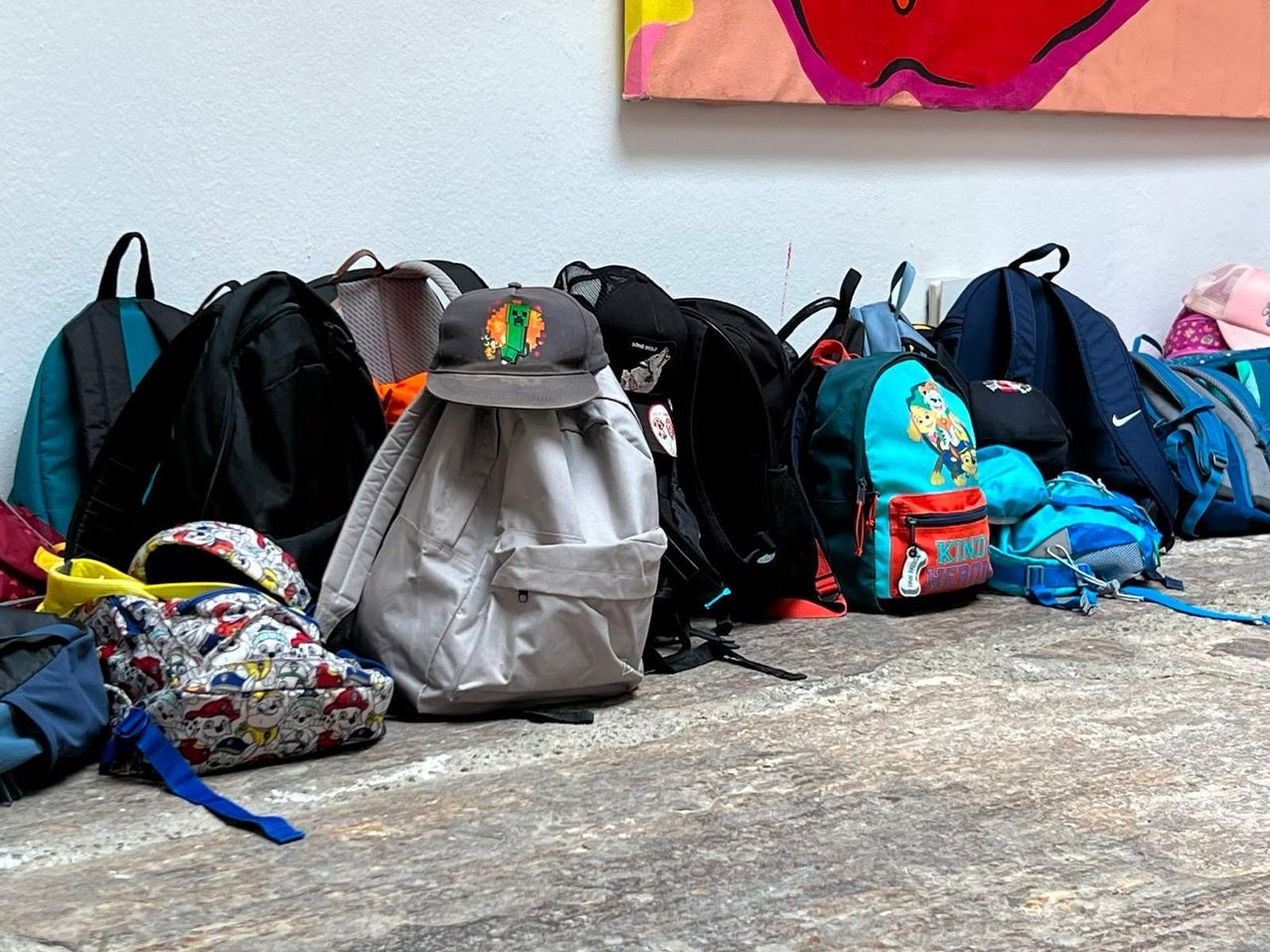 Eine Sammlung von Rucksäcken und Mützen ist vor einer farbenfrohen Wand auf einem Holzboden angeordnet. Ein grauer Rucksack mit einem Minecraft-Hut sticht unter ihnen hervor.