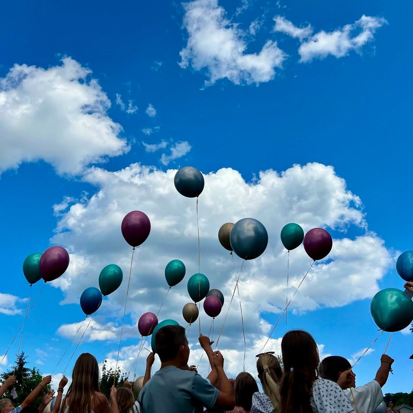 Eine Gruppe von Menschen lässt Ballons in einem blauen Himmel mit verstreuten Wolken frei. Die Ballons haben verschiedene Farben und Größen und steigen über die Menge auf.