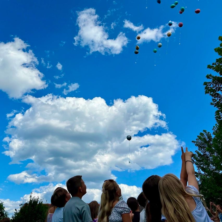 Eine Gruppe von Menschen beobachtet bunte Ballons, die in einem blauen Himmel mit weißen Wolken schweben. Ein Ballon ist in der Mitte, andere schweben herum.