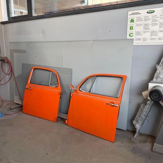 Zwei orangefarbene Autotüren sind an einer grauen Wand ausgestellt. Ein Fenster und ein Poster mit Buchstaben sind darüber. Ein roter Schlauch ist links.