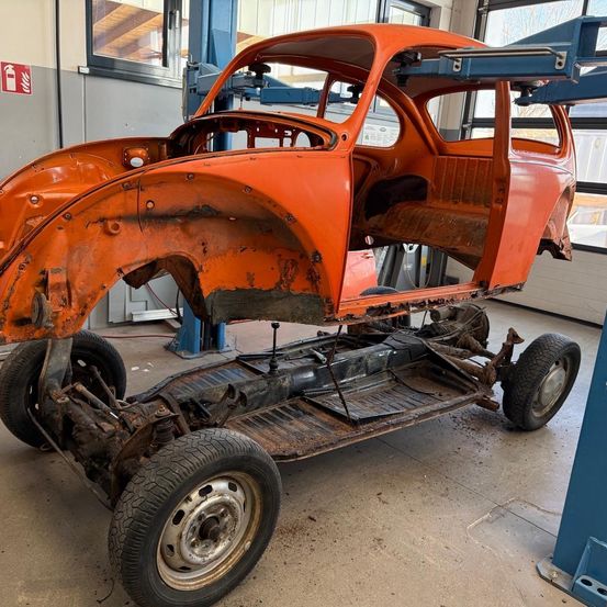 Ein alter orangefarbener Volkswagen Käfer ist in einer Werkstatt, ohne Karosserie, und wird von einem blauen Wagenheber angehoben.