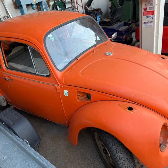 Ein alter orangefarbener VW Käfer mit beschädigtem vorderen Stoßfänger und einer Ersatzbereifung an der Seite steht in einer Garage.
