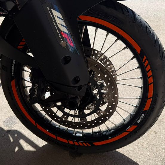 Das Vorderrad eines Motorrads zeigt einen silbernen Bremsscheiben und einen schwarzen Reifen mit orangefarbenem Felgenband. Das Rad ist am Gabelbaum des Motorrads befestigt.