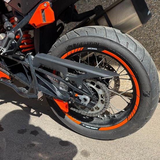 Das Hinterrad eines Motorrads zeigt einen orangefarbenen Felgenrand mit schwarzem Reifen. Die Kette und der Kettenritzel des Motorrads sind sichtbar.