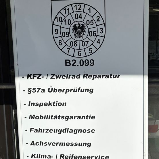Ein Schild zeigt die Dienstleistungen einer deutschen Autowerkstatt an. Es beinhaltet KFZ-Zweirad-Reparatur, 57a Überprüfung, Inspektion, Mobilitätsgarantie, Fahrzeugdiagnose, Achsvermessung und Klima-Reifenservice.