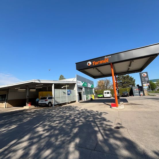 Eine Tankstelle mit Vordach zeigt ein Schild für Quick Turmöl. Ein weißer Wagen steht unter dem Vordach, und andere Fahrzeuge sind in der Nähe. Der Bereich ist gut von natürlichem Sonnenlicht beleuchtet.