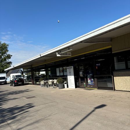 Ein Ladengeschäft mit Glastüren und -fenstern. Ein weißes Schild mit 'EINGANG' auf dem Dach. Stühle und ein Tisch sind draußen aufgestellt. Autos und Lieferwagen geparkt in der Nähe. Bäume in der Ferne.