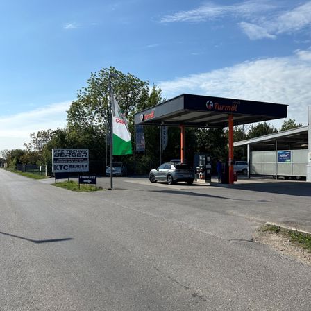 Eine leere Tankstelle mit einem grauen Auto, das an der Zapfsäule geparkt ist. Die Tankstelle hat ein Dach und mehrere Schilder, darunter eines mit der Aufschrift 'Einfahrt'.