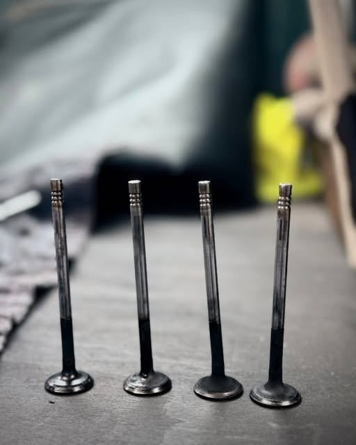 Vier metallische Zylinder sind auf einem Tisch aufgereiht und weisen Abnutzungsspuren auf. Im Hintergrund befinden sich verschwommene Stoffe und ein gelbes Objekt.