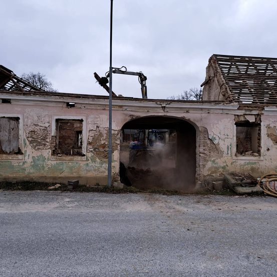 Ein verfallenes Gebäude mit zerbrochenen Fenstern und einem eingestürzten Dach wird von einem Baufahrzeug abgerissen, wobei eine Staubwolke entsteht.