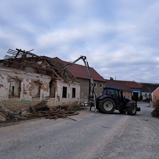Ein Traktor mit einem Kran steht vor einem beschädigten Haus, das ein halbes zerstörtes Dach und einen Schutthaufen hat.