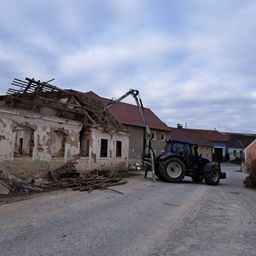 Ein Traktor mit einem Kran steht vor einem beschädigten Haus, das ein halbes zerstörtes Dach und einen Schutthaufen hat.