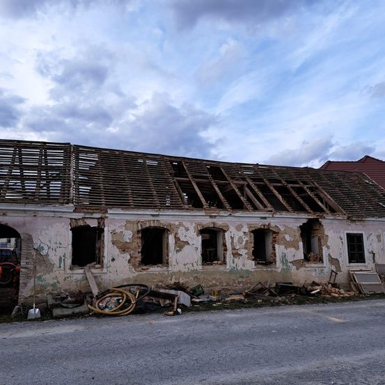 Ein altes, baufälliges Gebäude mit einem beschädigten Dach und zerbrochenen Fenstern. Schutt und Materialien sind um das Gebäude herum verstreut.