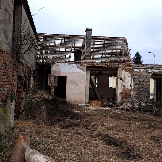 Ein altes Backsteingebäude ist teilweise abgerissen, wobei eine Holzkonstruktion das Dach stützt. Das Gebiet ist mit Schutt und trockenem Gras gefüllt.