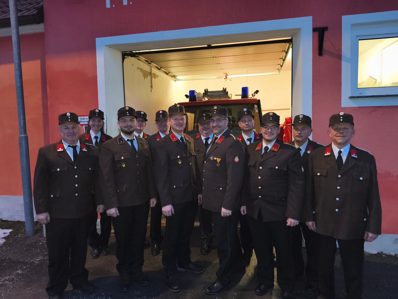 A group of firefighters dressed in uniforms, standing in front of a fire station. A fire engine is visible in the background through the open garage door.