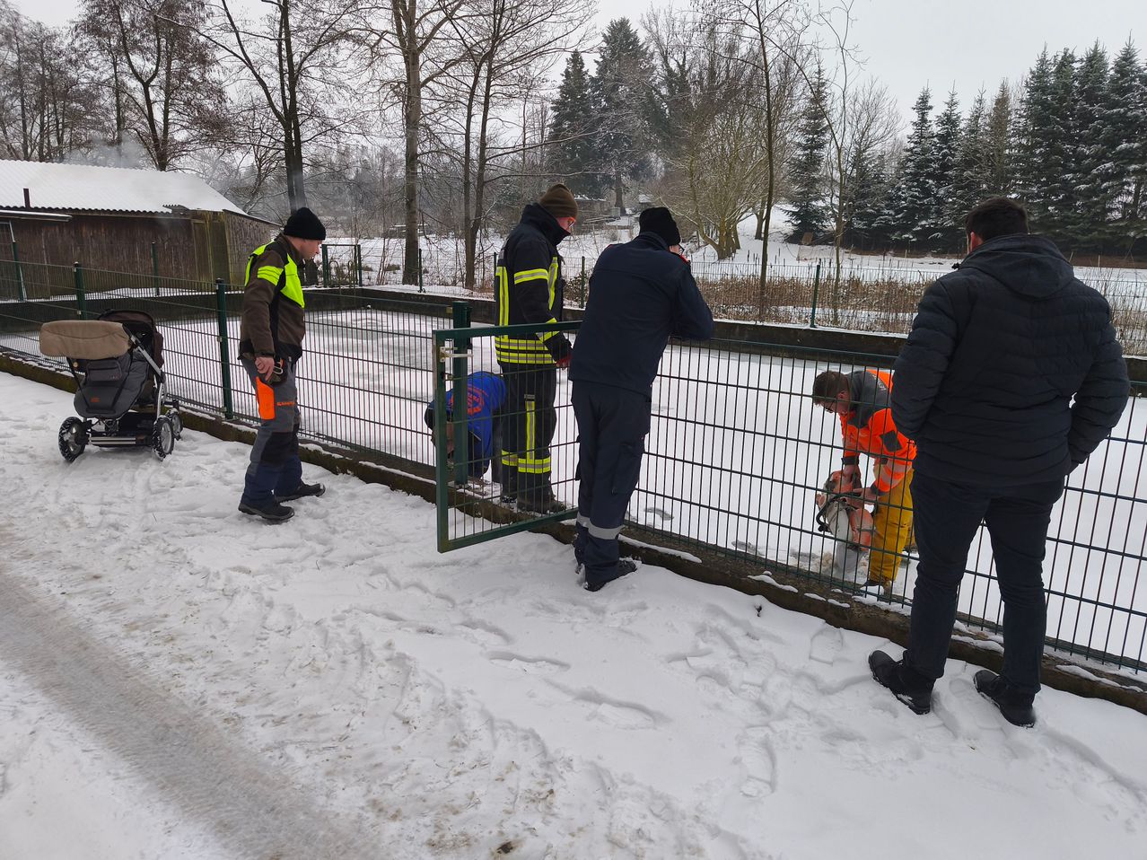 Mehrere Arbeiter in orangefarbenen Uniformen arbeiten in einem verschneiten Bereich, einer reinigt den Zaun mit einem Schlauch.