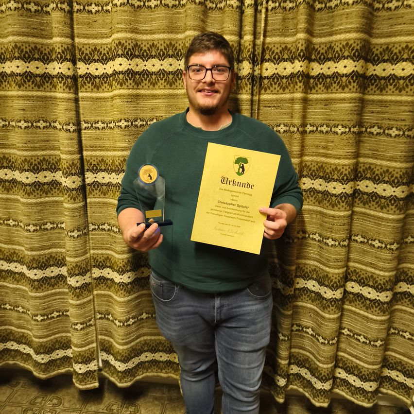 Ein Mann mit Brille und einem grünen Shirt hält eine Urkunde und einen Pokal mit Baumlogo. Er steht vor einem gemusterten Vorhang.