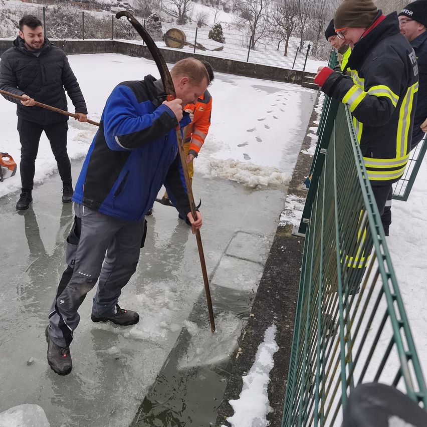 Eine Gruppe von Menschen räumt Eis von einer gefrorenen Oberfläche mit Stöcken. Ein Mann lehnt sich über eine Stange, während andere von einem Zaun aus zusehen.