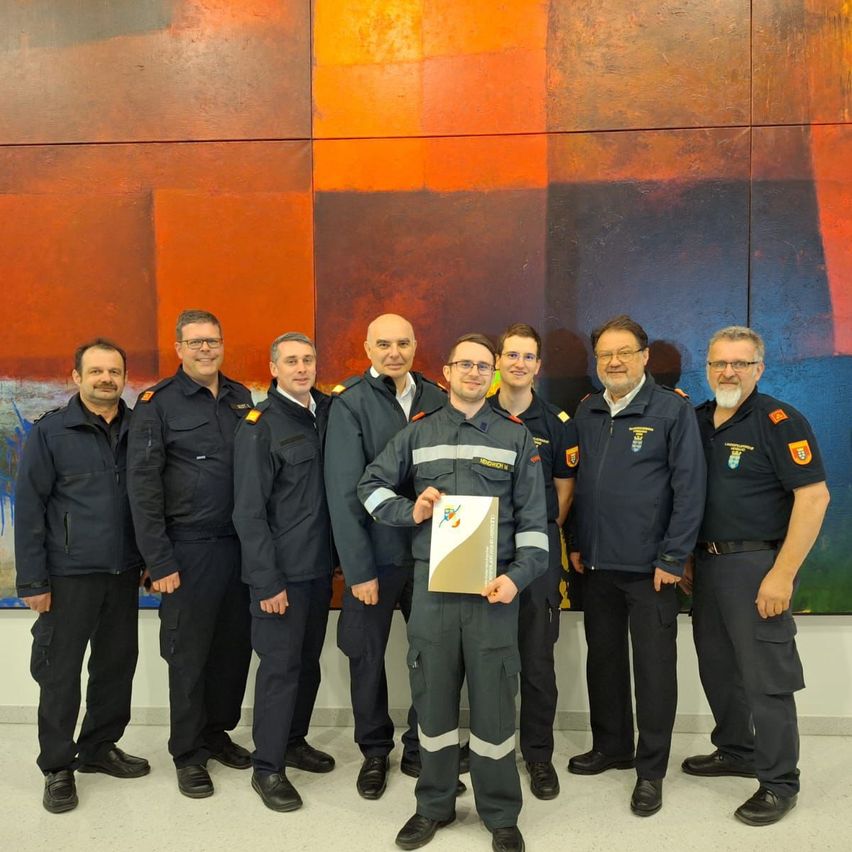 Eine Gruppe von Feuerwehrleuten in Uniformen posiert für ein Foto vor einer großen farbenfrohen Wand. Einer von ihnen hält ein Zertifikat.