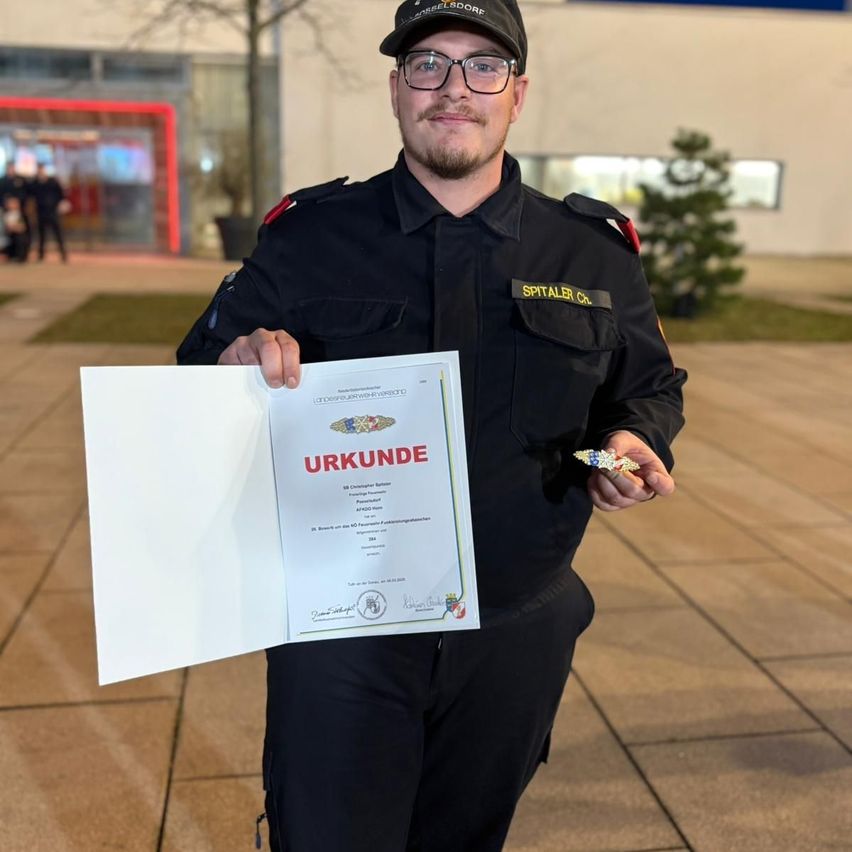Ein Polizist hält ein Zertifikat mit dem Wort 'URKUNDE' darauf. Er trägt eine Uniform und hält eine Medaille. Hinter ihm steht ein Gebäude mit einer roten Leuchte und einem kleinen Baum.