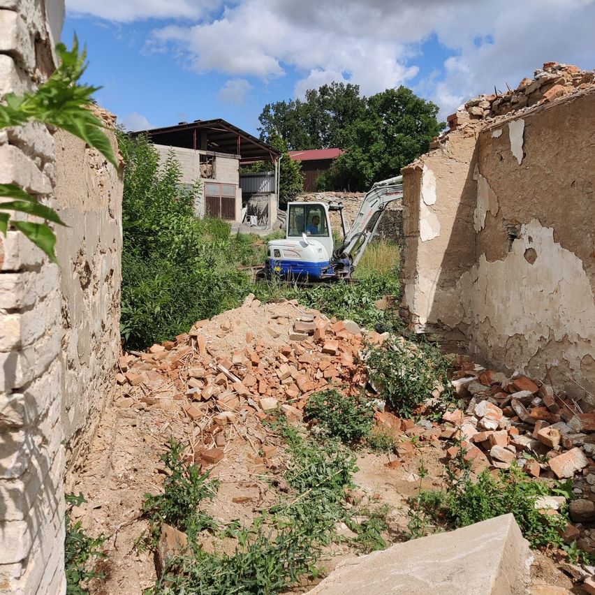 Ein Abrissplatz mit einem weißen und blauen Bagger, der Schutt entfernt. Dahinter ein teilweise abgerissenes Gebäude und ein Haus im Bau.