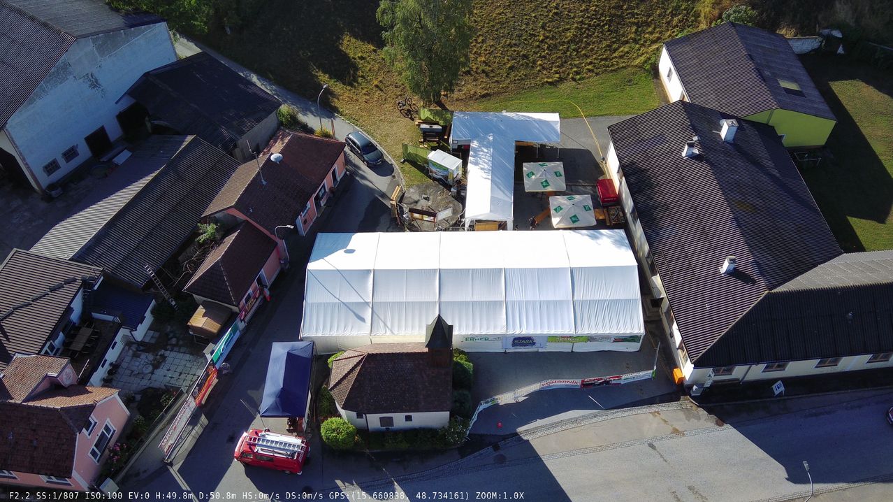 An aerial view of a rural area with several buildings, a large white tent, and a red fire truck parked nearby. The area is surrounded by greenery and has a small road.