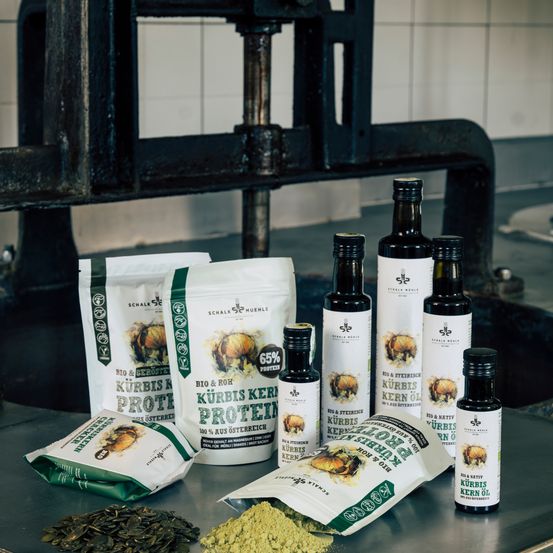 Verschiedene Flaschen mit Kürbiskernöl, zusammen mit Tüten mit Kürbiskernprotein und -pulver, sind auf einem Tisch in einer Küche angeordnet.