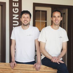Zwei Männer in weißen T-Shirts und Jeans posieren neben einer Holzbank, lächelnd, mit einem Gebäude und einem Glasfenster im Hintergrund.